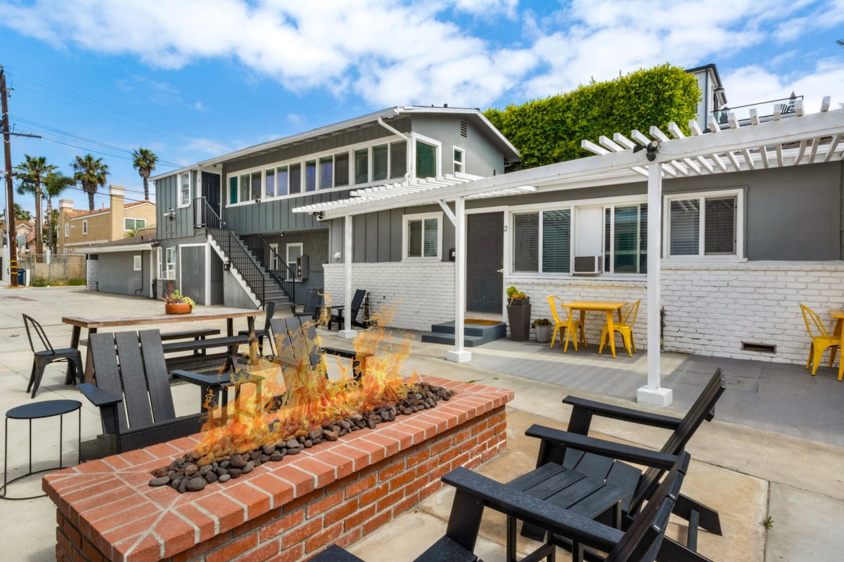 An outdoor communal area features a red brick fire pit surrounded by black chairs. Yellow patio chairs are positioned nearby under a white pergola. The background showcases a multi-level building with large windows and green landscaping, creating a well-lit and inviting space.