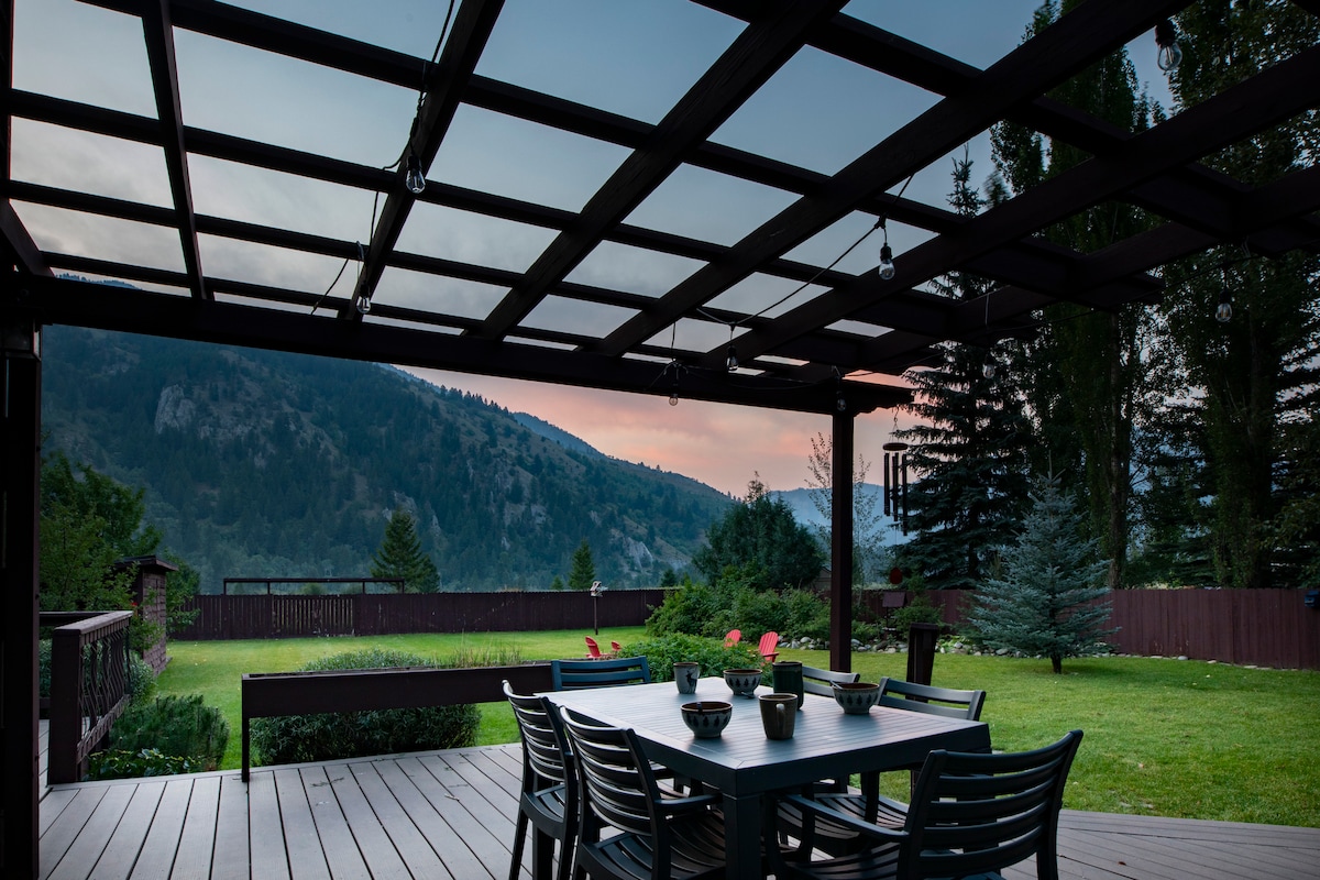An outdoor dining space is presented underneath a structured pergola, overlooking expansive green lawn and distant mountain views. A large table with multiple chairs is positioned centrally, complemented by decorative bowls. Soft evening light creates a serene atmosphere, enhancing the natural beauty of the surroundings.