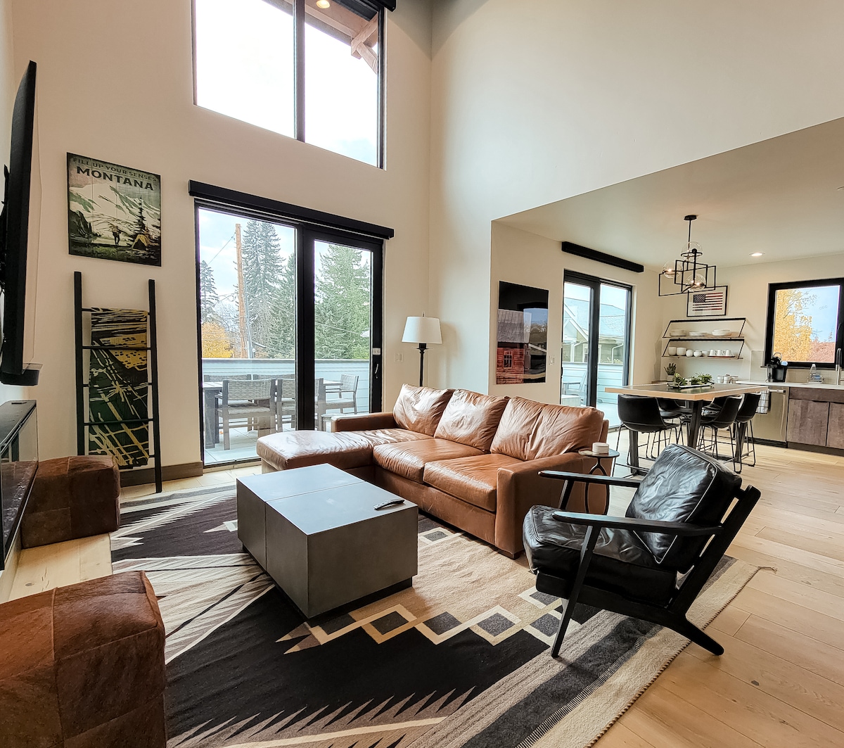 A spacious living area features large windows allowing natural light to fill the room. A comfortable leather sofa and an armchair are arranged around a minimalist coffee table. A stylish dining space is visible in the background, with outdoor access leading to a balcony.