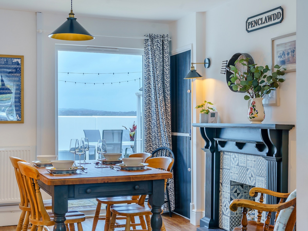 The living and dining area features a wooden dining table surrounded by chairs, set against a backdrop of coastal views through glass doors. A decorative fireplace and wall art complete the space, while soft lighting adds to the inviting ambiance.