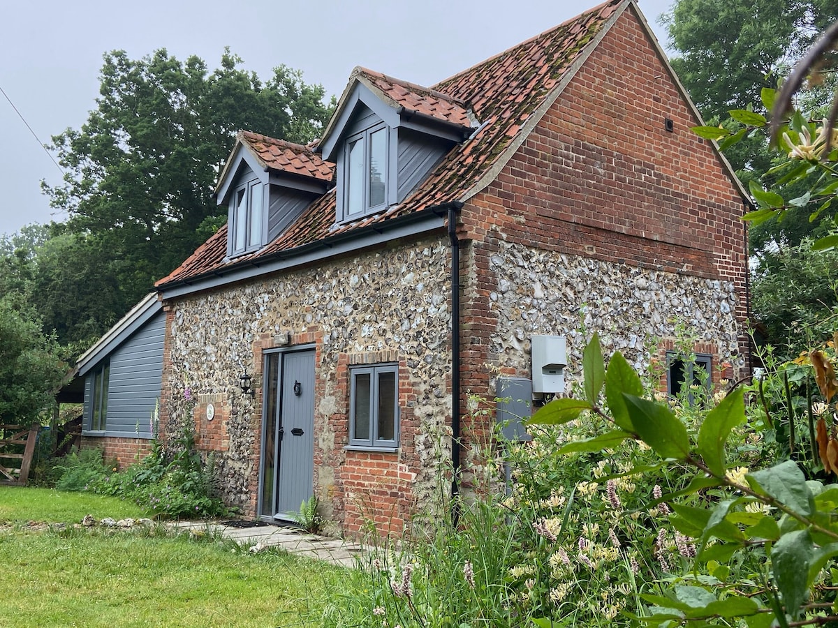 A charming two-story cottage features a stone exterior coupled with red brick detailing. Grey shutters accentuate the windows above, while a blue door welcomes guests. Lush greenery and flowering plants surround the property, enhancing the tranquil outdoor setting.
