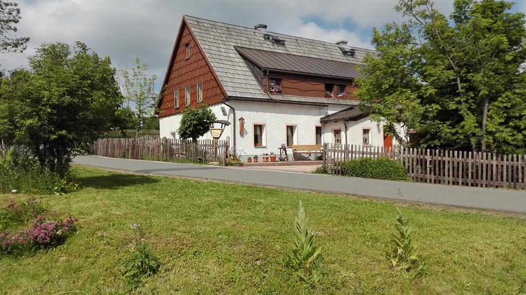 A traditional two-story building with wooden features is set against a cloudy sky. The exterior is predominantly white, complemented by brown accents. Lush greenery surrounds the property, with a well-maintained garden and a picket fence along the path. Outdoor seating is visible near the entrance.