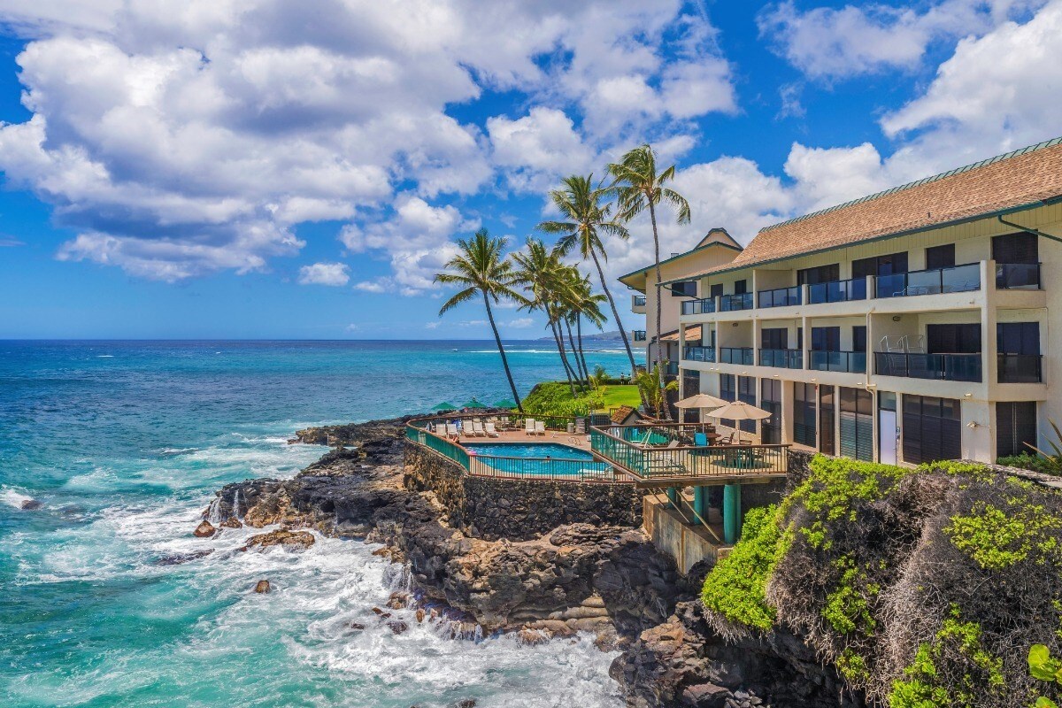 An oceanfront view showcases a multi-story building with balconies overlooking the turquoise water. Palm trees line the coastline, and a pool area is visible on the rocky shore, offering direct access to the sea amid vibrant blue skies with scattered clouds.