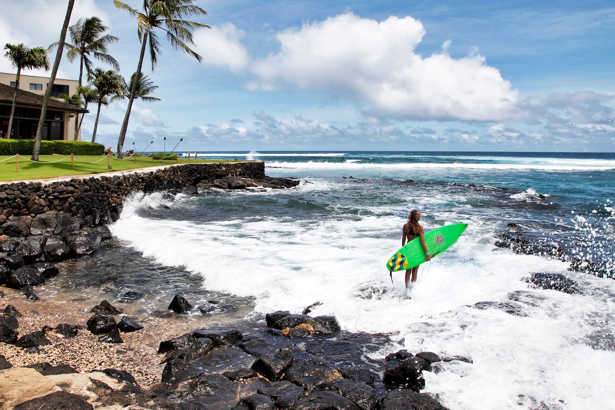 A surfer stands at the water's edge, holding a bright green surfboard. Waves crash against the rocky shore, while palm trees are visible in the background under a mostly cloudy sky. The ocean stretches out into the distance, inviting exploration.