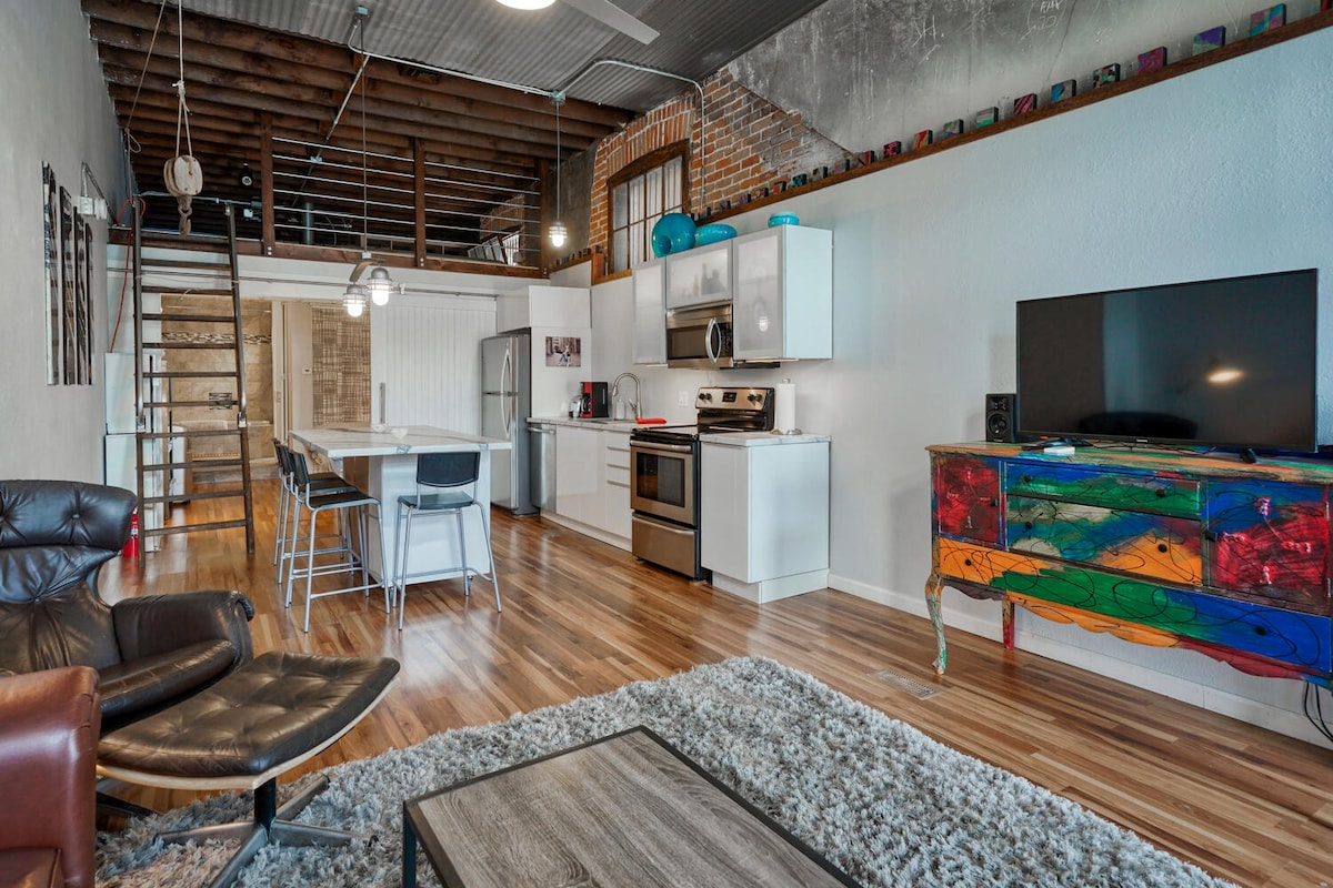 An open living space is highlighted by polished wooden floors and high ceilings. A fully equipped kitchen features modern appliances, while a dining table with stools stands nearby. A large colorful cabinet offers visual interest, and a comfortable seating area includes two chairs and a coffee table.