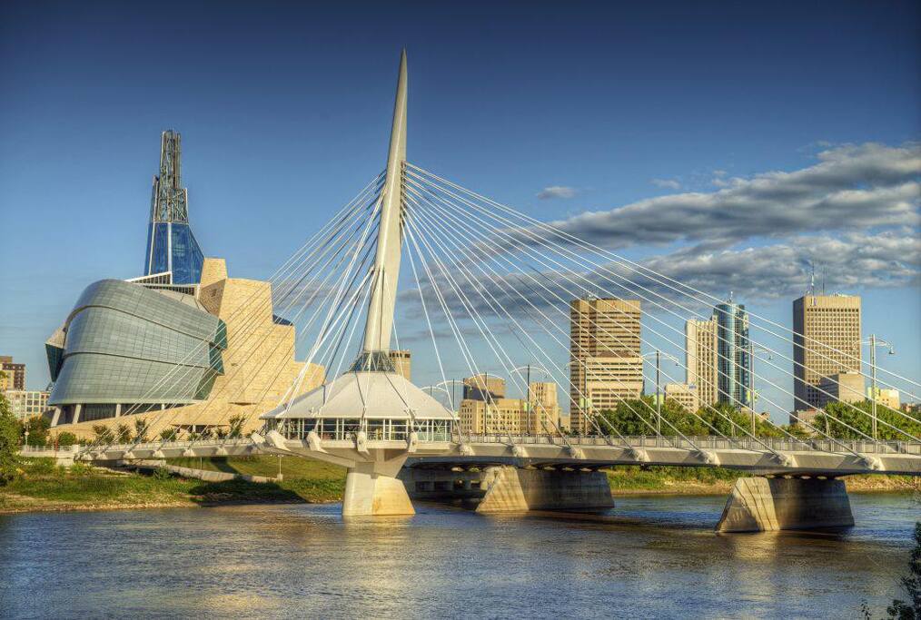 A modern bridge is shown spanning a serene river, connecting the two banks. The skyline features a mix of contemporary buildings with sleek lines and varying heights. A distinctive, cable-stayed structure stands prominently, while the water reflects the clear blue sky and soft clouds.