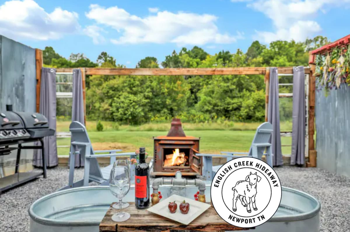 An outdoor patio area features a rustic firepit surrounded by seating. Two glasses and a bottle of wine accompany a small plate of chocolate-covered strawberries, suggesting an inviting space for relaxation. Grills are positioned at the edge, with lush greenery visible in the background.