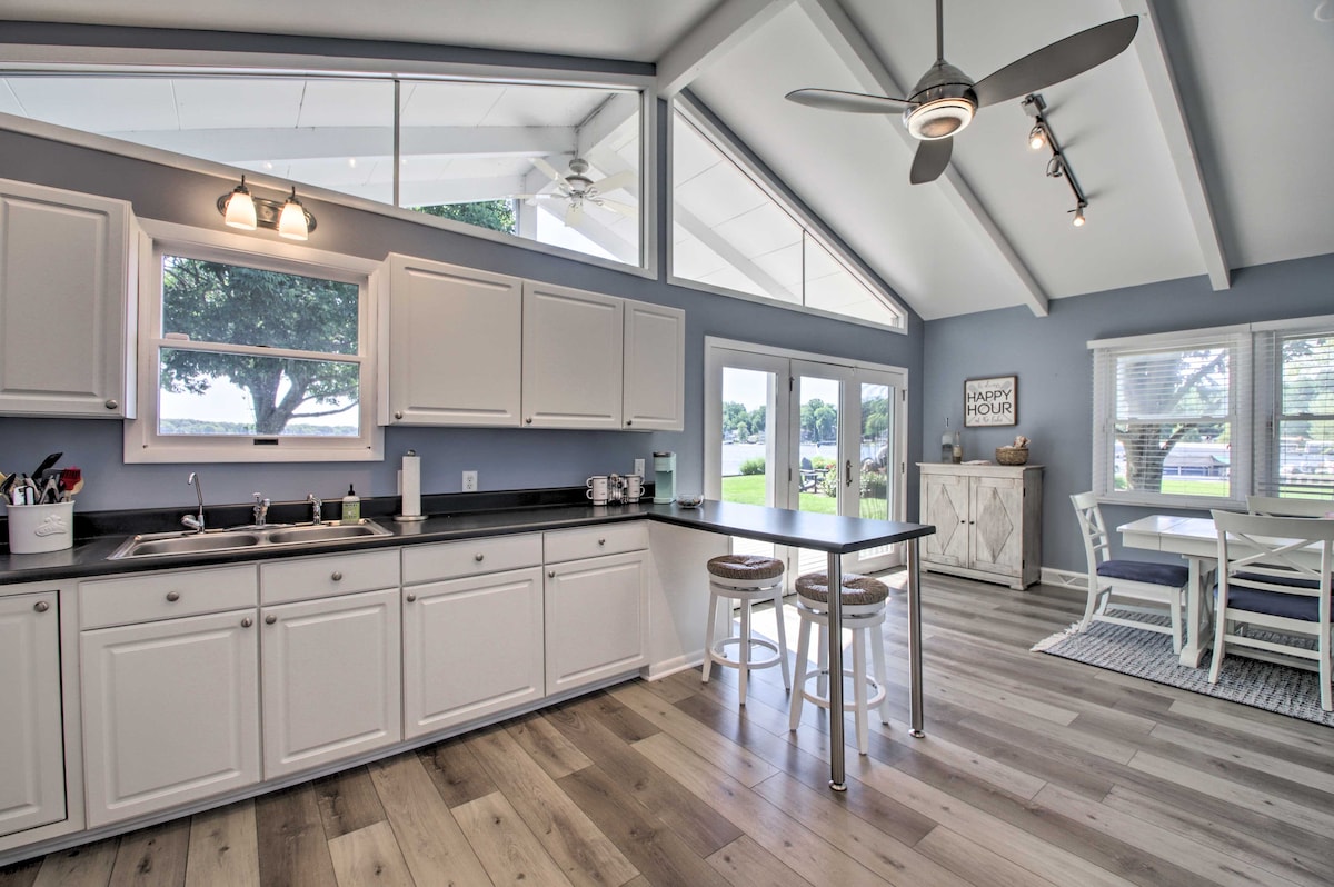 The kitchen area features white cabinetry and a dark countertop, framed by large windows that allow natural light to fill the space. A dining table and additional seating are visible, with an open floor plan leading to a light-filled living area.