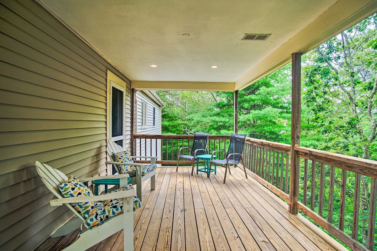 A furnished deck offers two comfortable chairs and a small table, framed by wooden railings. The space is surrounded by lush green trees, enhancing a serene outdoor environment. The deck features wooden flooring, providing a cozy area to enjoy the natural surroundings.