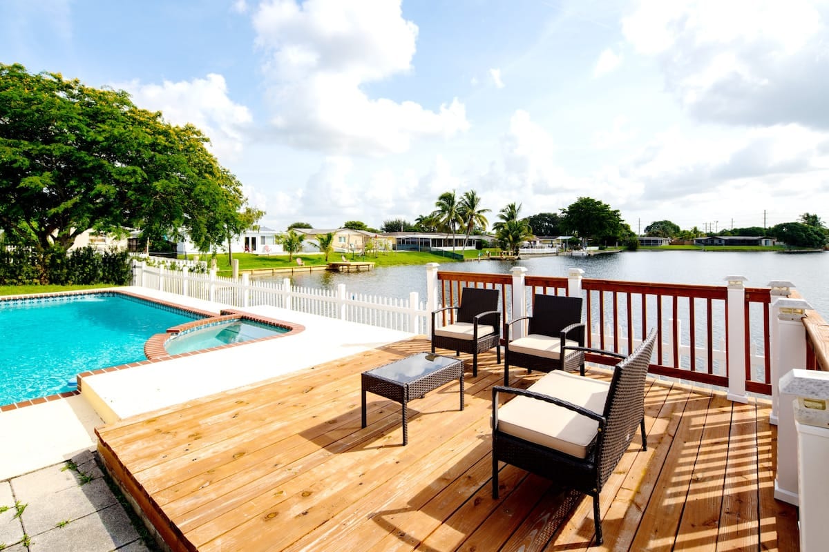 A wooden deck features a heated swimming pool and seating area with chairs and a small table. The scenic lake is visible in the background, framed by lush greenery and palm trees. Bright clouds float across the clear blue sky, enhancing the outdoor ambiance.