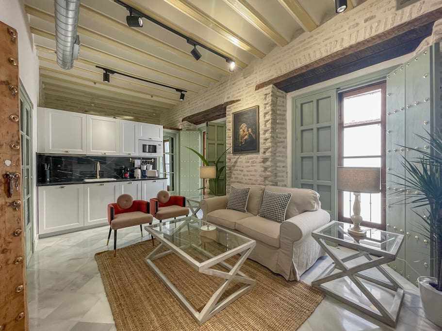 A cozy living area features a light-colored sofa and two modern red chairs surrounding a glass coffee table. The kitchen area is visible, with sleek cabinetry and appliances. Natural light floods in through open green shutters, illuminating the whitewashed brick walls.