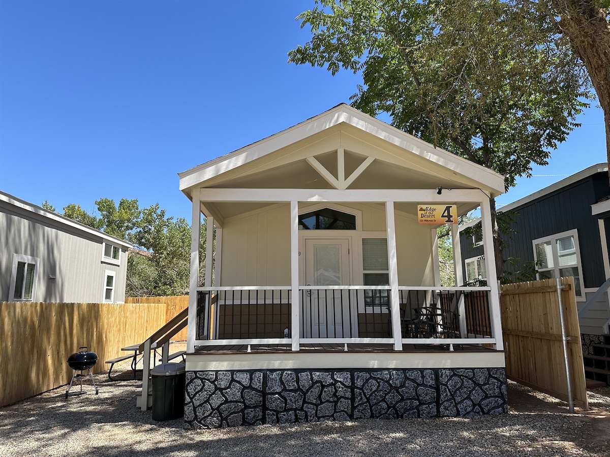 The cabin's front exterior is framed by a porch with a sloped roof and wooden railing. A sign labeled 'Cabin #4' hangs at the entrance. A charcoal grill and a trash bin are positioned beside the porch, with gravel underfoot and trees providing shade.