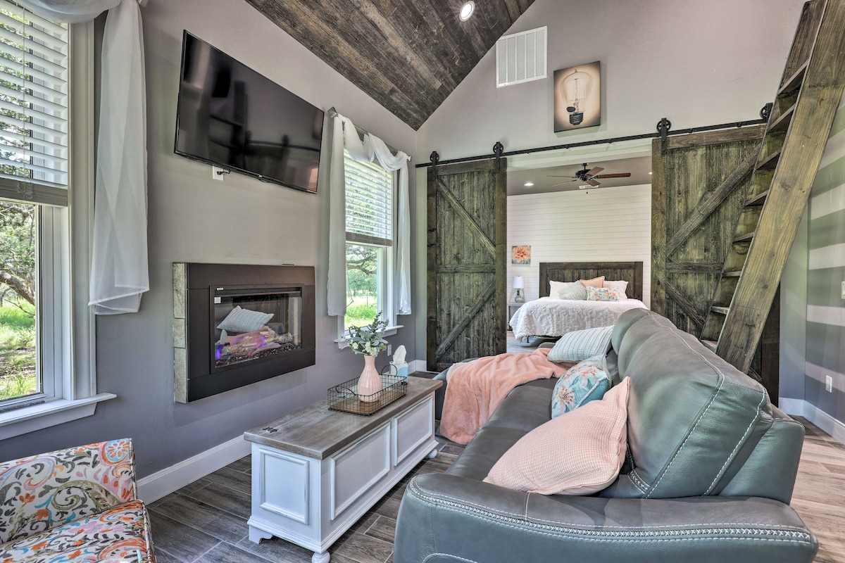 An inviting living area showcases a comfortable sofa with decorative pillows positioned near a modern electric fireplace. A Smart TV is mounted above, while large windows allow natural light to illuminate the space. Wooden accents and a loft area add a rustic charm.