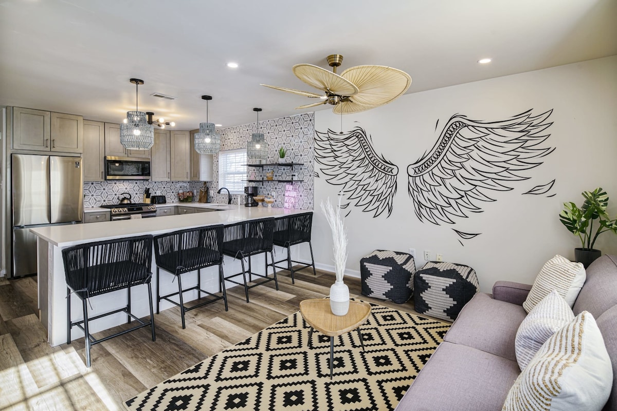 An open living area features a stylish kitchen with modern appliances and bar seating. A unique winged mural adorns the wall, complemented by a patterned rug and comfortable seating. Decorative plants add a fresh touch to the inviting space.