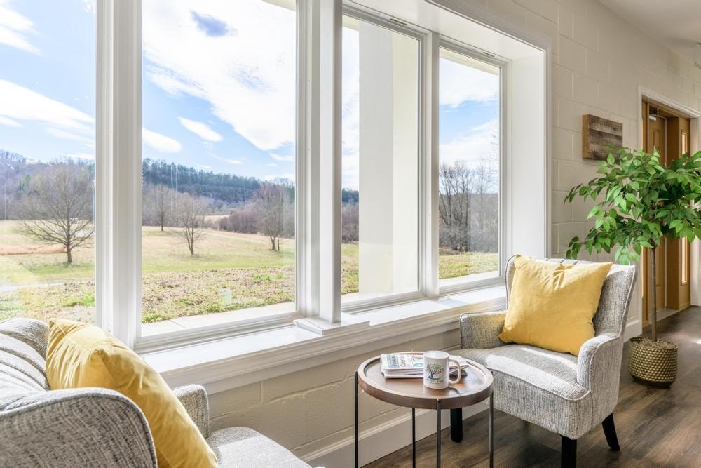 A cozy seating area is positioned beside large windows, inviting natural light and offering views of open fields and distant hills. Two armchairs are accompanied by a small side table, which is set with a mug and a magazine, creating a peaceful nook for relaxation.