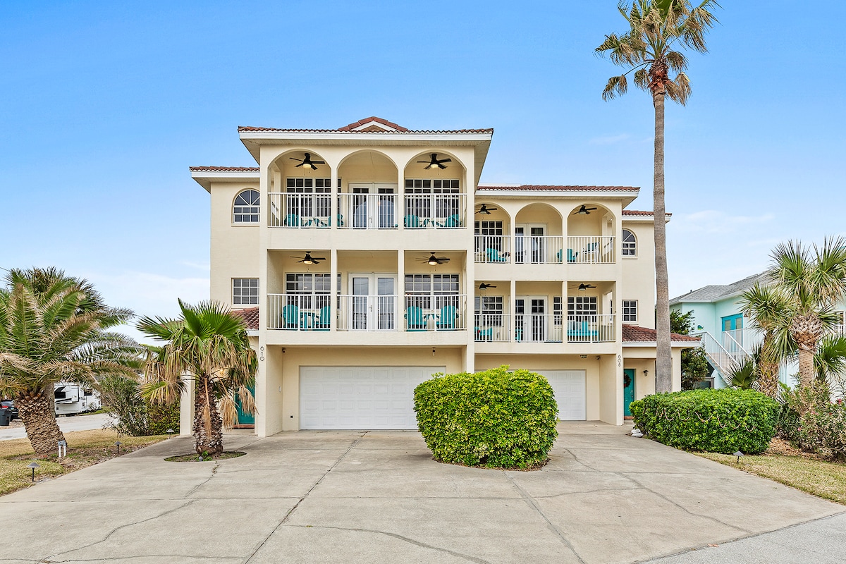 A three-story townhome is presented, featuring multiple balconies and large windows. The facade is adorned with arched details and ceiling fans. Surrounding greenery includes palm trees and neatly manicured shrubs, while a spacious driveway leads to the garage area below.