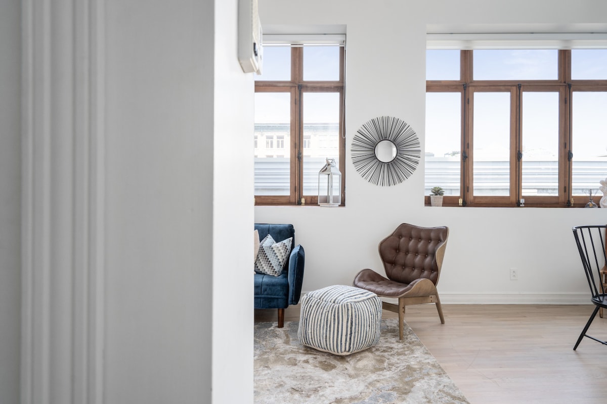 A bright corner of the apartment features large windows allowing natural light to fill the space. A cozy seating area includes a blue armchair, a stylish leather chair, and a round pouf. Decorative elements, such as a sunburst mirror and a soft rug, enhance the ambiance.