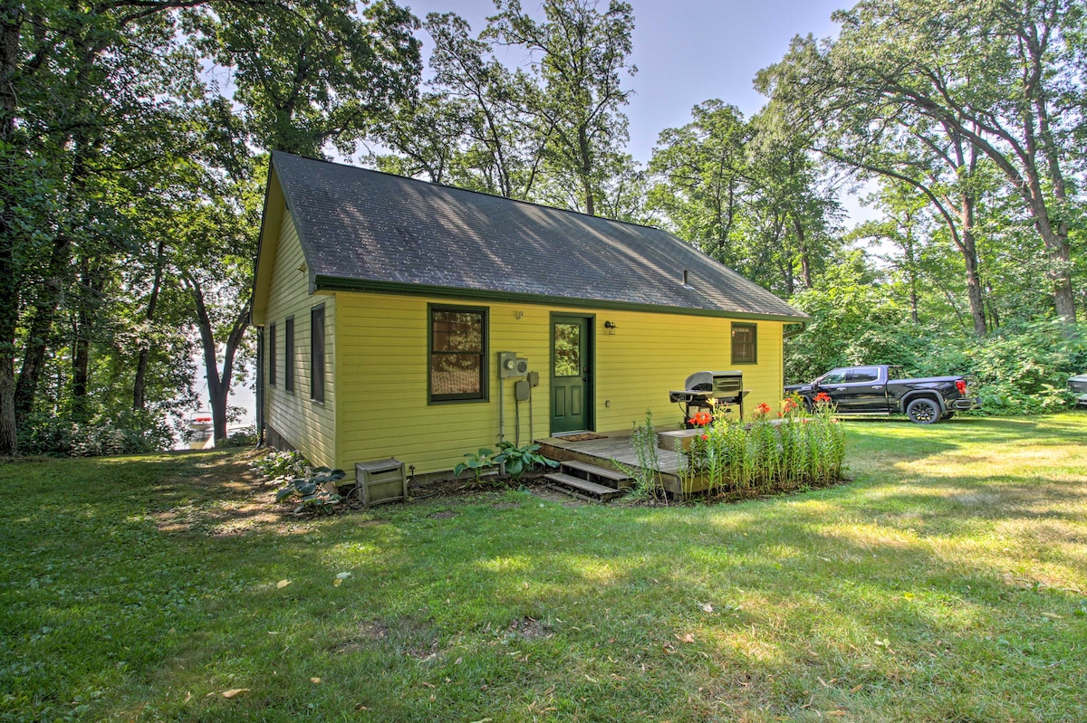 A yellow cabin is nestled among trees, revealing a deck with a gas grill and flower beds in the foreground. A grassy yard extends towards a nearby body of water, with a vehicle parked at the side of the cabin.