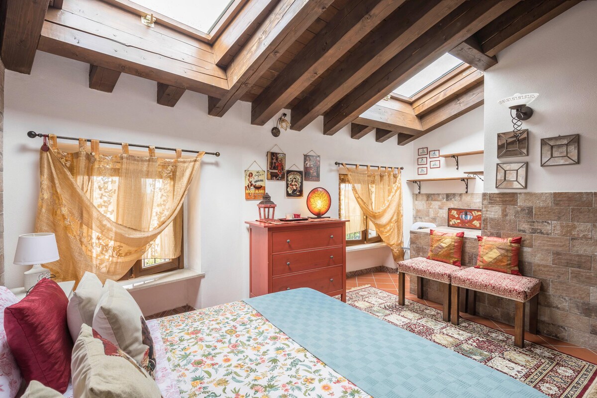 A comfortable bedroom features a spacious bed adorned with a patterned blanket. Natural light filters through skylights and window drapes, illuminating the rustic wooden beams overhead. A dresser and seating area are visible, complemented by decorative wall art and a warm-toned lamp.