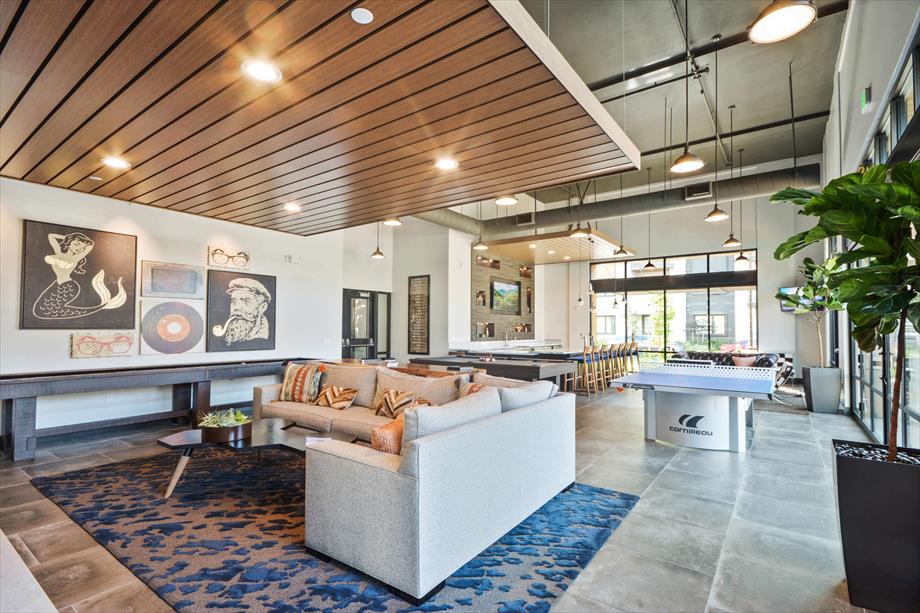 A spacious lounge area is highlighted by a modern design featuring a comfortable sectional sofa and a decorative area rug. Large windows allow natural light to fill the space, while artwork adorns the walls. A communal dining area is visible in the background.