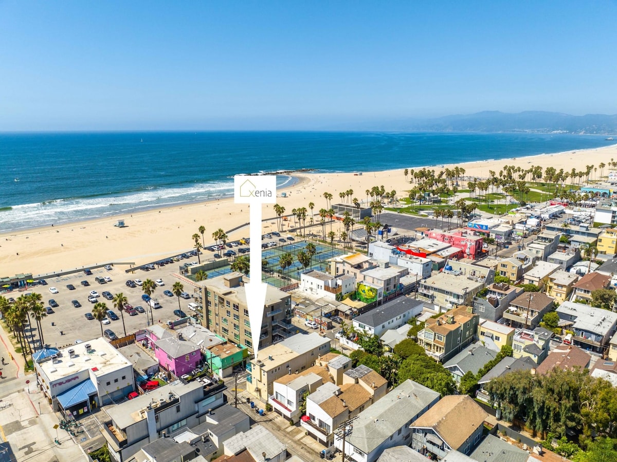 An aerial view showcases the beachfront location of the property, with the ocean in view and the Venice Beach Boardwalk visible in the distance. The vibrant colors of local shops and palm trees create a lively coastal scene.