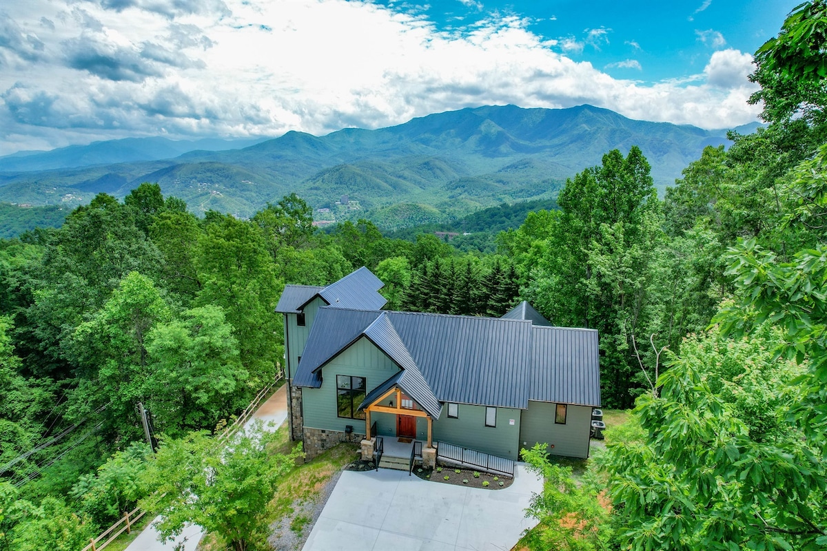The exterior of a newly built luxury home is showcased against a backdrop of rolling mountains. Lush green trees surround the property, with a large, flat driveway leading to the entrance. The home features a modern design with a dark-colored roof and warm wooden accents.