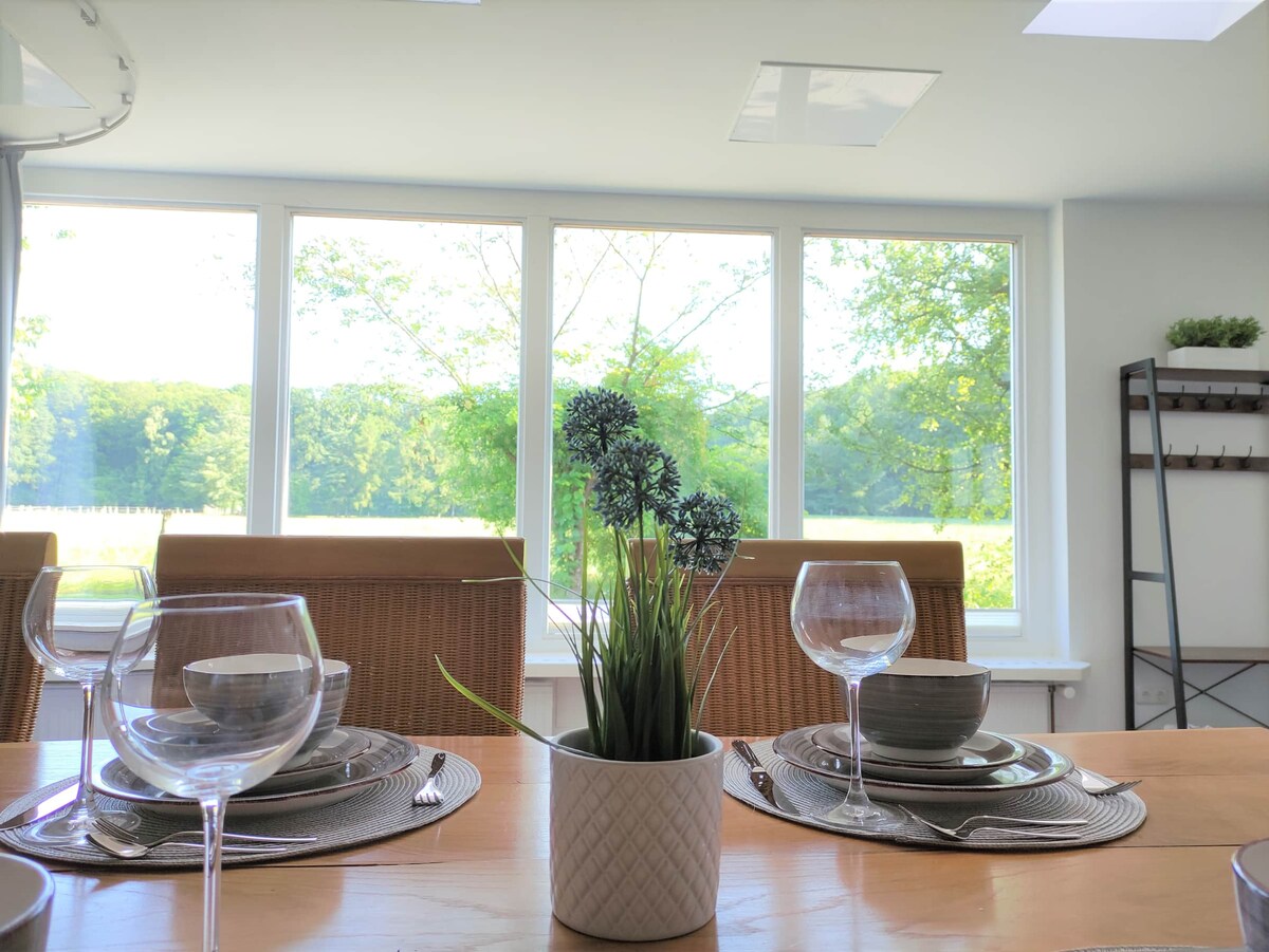 A dining area is arranged with elegant table settings, including plates, cups, and wine glasses. A small potted plant sits at the center of the table. Large windows provide a view of lush greenery outside, inviting natural light into the space.