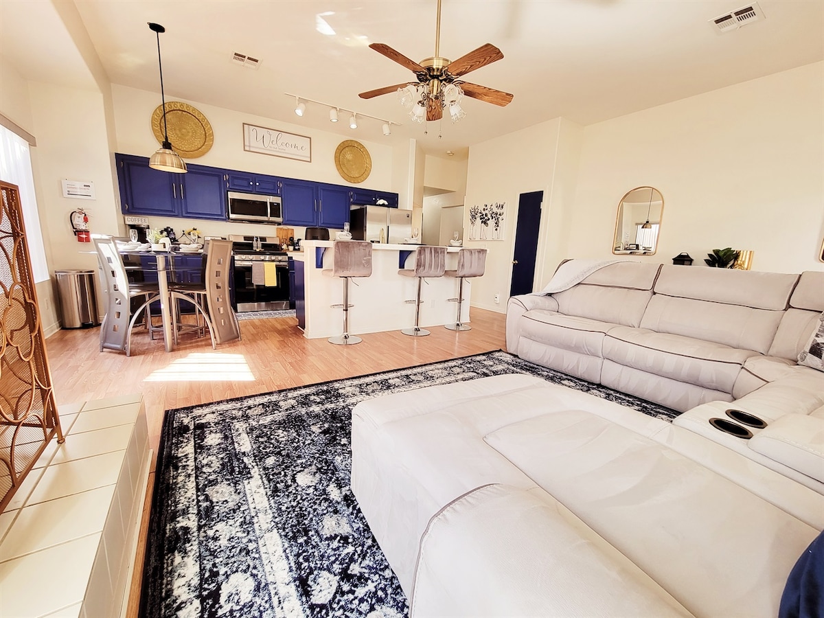 A spacious living area is highlighted by a light-colored sectional sofa and a patterned area rug. The kitchen features dark blue cabinetry and modern appliances, with bar seating visible. Natural light fills the room through windows, creating an inviting atmosphere.
