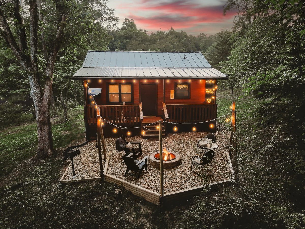 The cabin exterior is surrounded by lush greenery and features a welcoming porch adorned with string lights. An outdoor fire pit is present, accompanied by several seating options, including rustic chairs. A charcoal grill is positioned nearby, enhancing the outdoor recreational space.