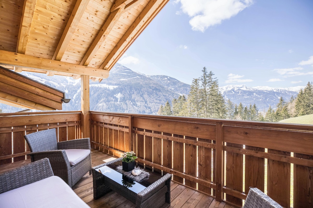 A balcony is furnished with comfortable seating and a small table, surrounded by wooden railings. Spacious views of the mountains and pine trees can be seen in the background, under a clear blue sky with a few clouds.