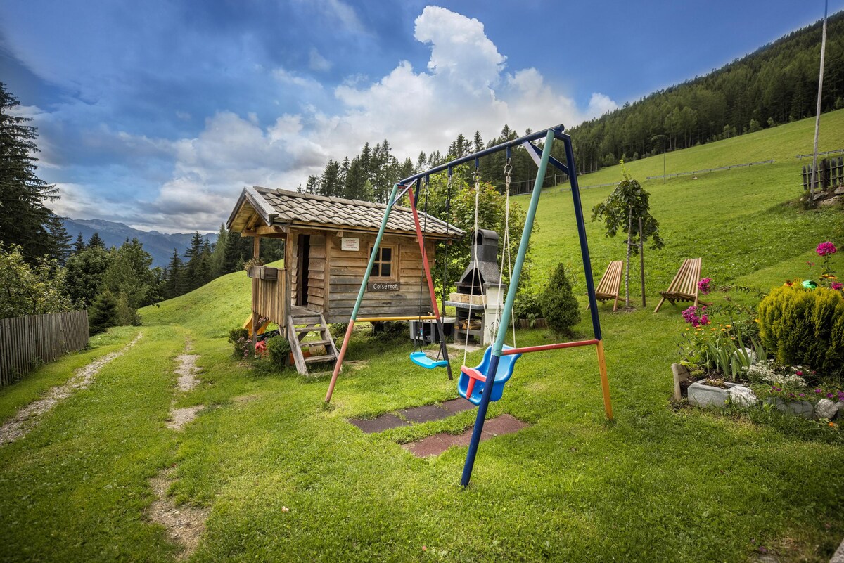 A grassy area features a wooden playhouse alongside a colorful swing set. Two lounge chairs are positioned nearby, surrounded by vibrant flowers. The backdrop showcases rolling hills and a partly cloudy sky, providing a serene setting for play and relaxation.