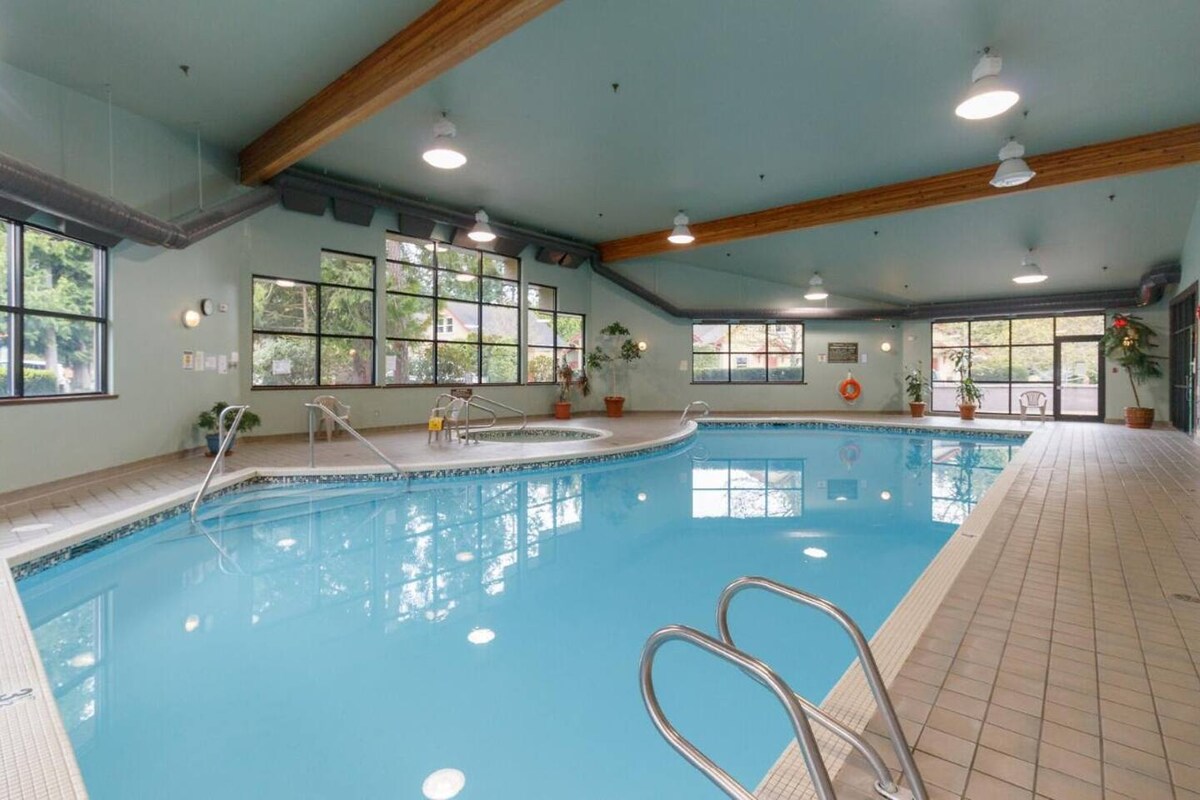 An indoor swimming pool is highlighted, featuring clear blue water and a tiled surround. Large windows provide ample natural light, offering views of the outside. Potted plants are situated throughout the area, creating a serene environment. Safety equipment is visible along the wall.