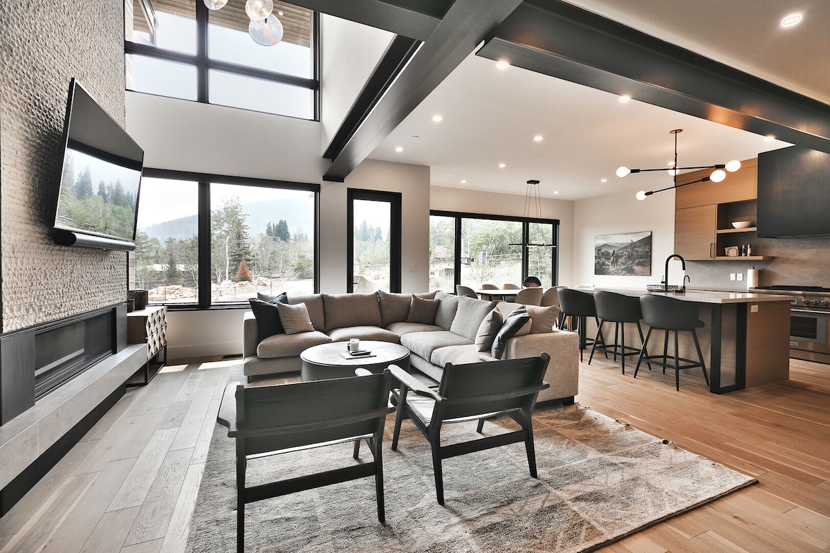 The open living area showcases a spacious sectional sofa, complemented by modern armchairs. Large windows provide natural light and picturesque views of the surrounding nature. A sleek television is mounted on the wall above a contemporary fireplace, while a dining space with stylish light fixtures is visible in the background.