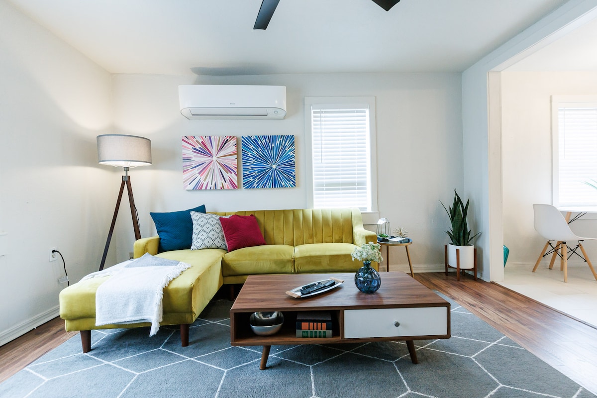 A modern living area features a bright yellow sectional sofa adorned with colorful pillows. A minimalist coffee table holds decorative items. Framed artwork hangs on the wall, complemented by natural light from nearby windows. A sleek floor lamp adds a warm touch to the space.
