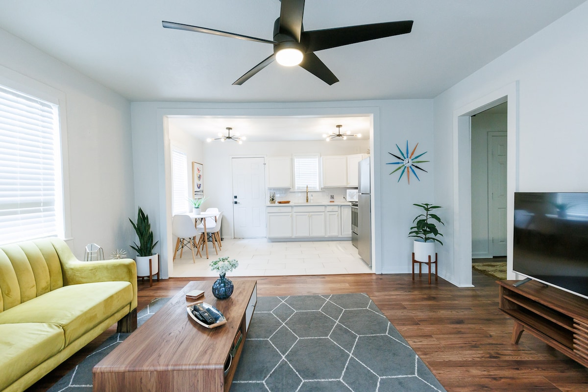 A modern living room is showcased with a vibrant yellow couch facing a sleek wooden coffee table. A geometric gray rug anchors the space, while large windows allow natural light to fill the area. The open layout transitions smoothly into a bright kitchen visible in the background.