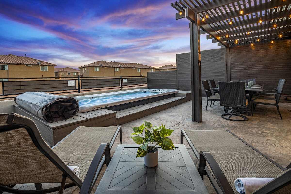 An outdoor space features a personal swim spa surrounded by wooden decking. Comfortable seating includes lounge chairs and a dining table, all beneath a structured pergola adorned with string lights. A potted plant adds a touch of greenery to the inviting atmosphere.