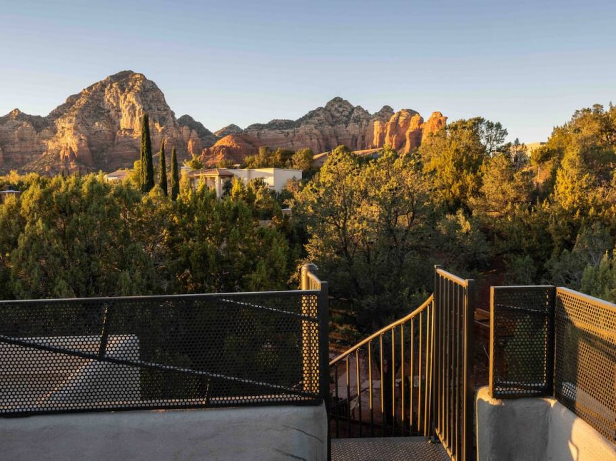 The rooftop deck provides expansive views of surrounding red rock formations and lush greenery. A winding metal staircase leads down from the deck, connecting to the lower levels of the property. The early morning light casts warm hues across the landscape.
