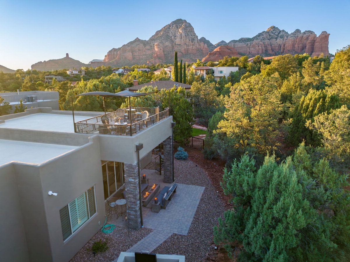 A modern home features a spacious outdoor area with seating and a fire pit, set against a backdrop of red rock formations. Lush greenery surrounds the property, and the rooftop deck provides stunning views of the scenic landscape.