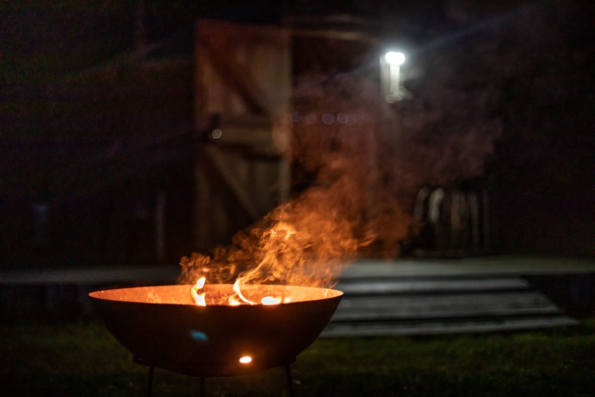 A fire basket burns brightly in the foreground, emitting wisps of smoke that dance in the night air. In the background, a softly lit structure is partially visible, with wooden doors creating a rustic ambiance.