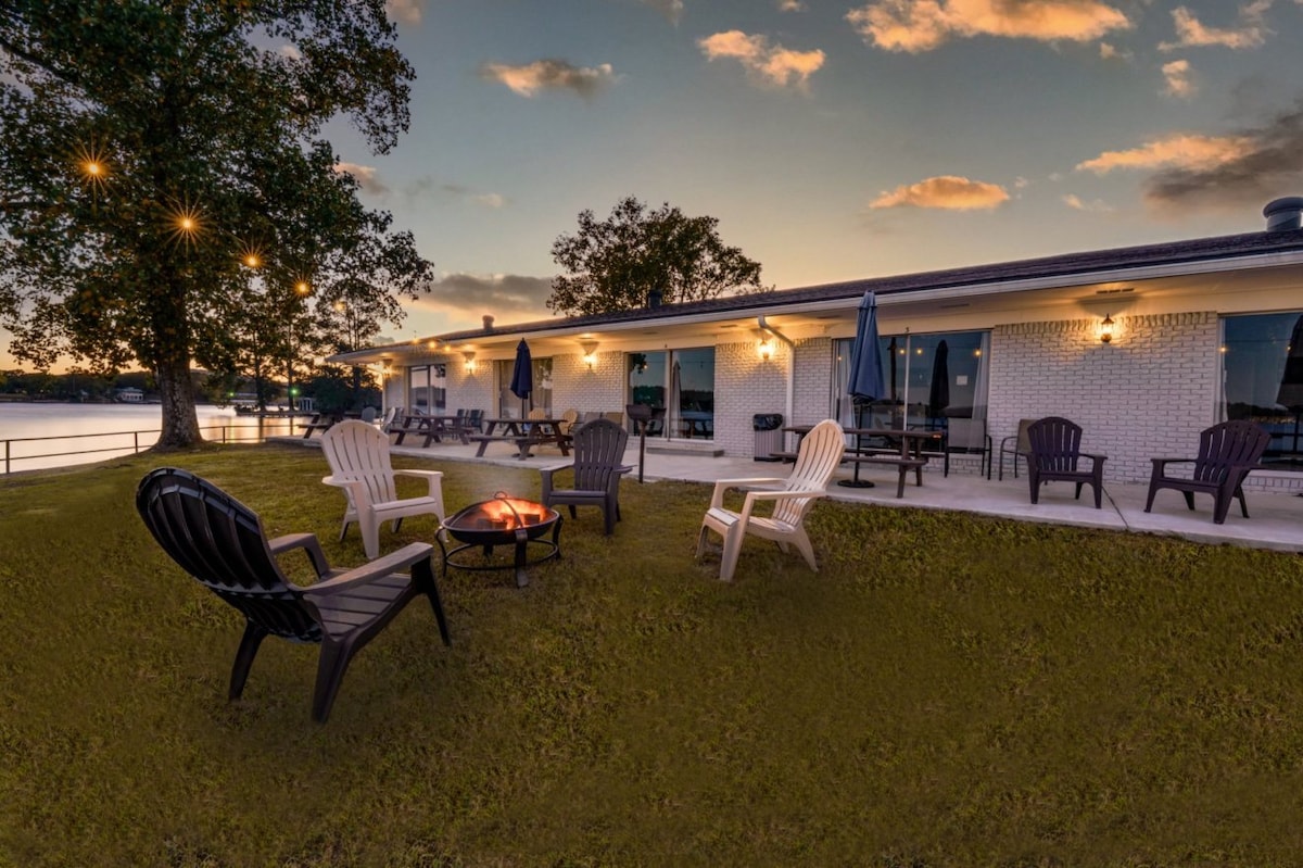 An outdoor seating area is depicted with several Adirondack chairs arranged around a fire pit. Soft lighting is provided by string lights above, and a serene view of the lake is visible in the background. The setting invites relaxation in a tranquil environment.