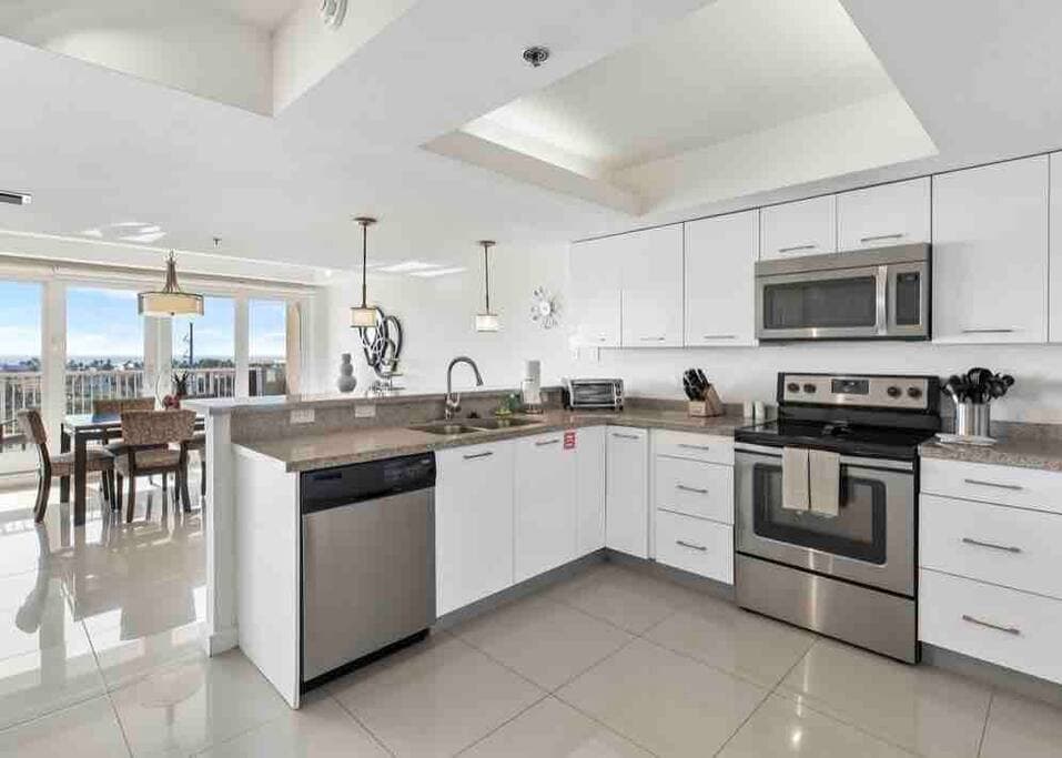 The modern kitchen features sleek white cabinetry and stainless steel appliances, including a dishwasher and oven. A large island provides additional counter space, while natural light illuminates the area through nearby windows. Dining space is visible in the background.