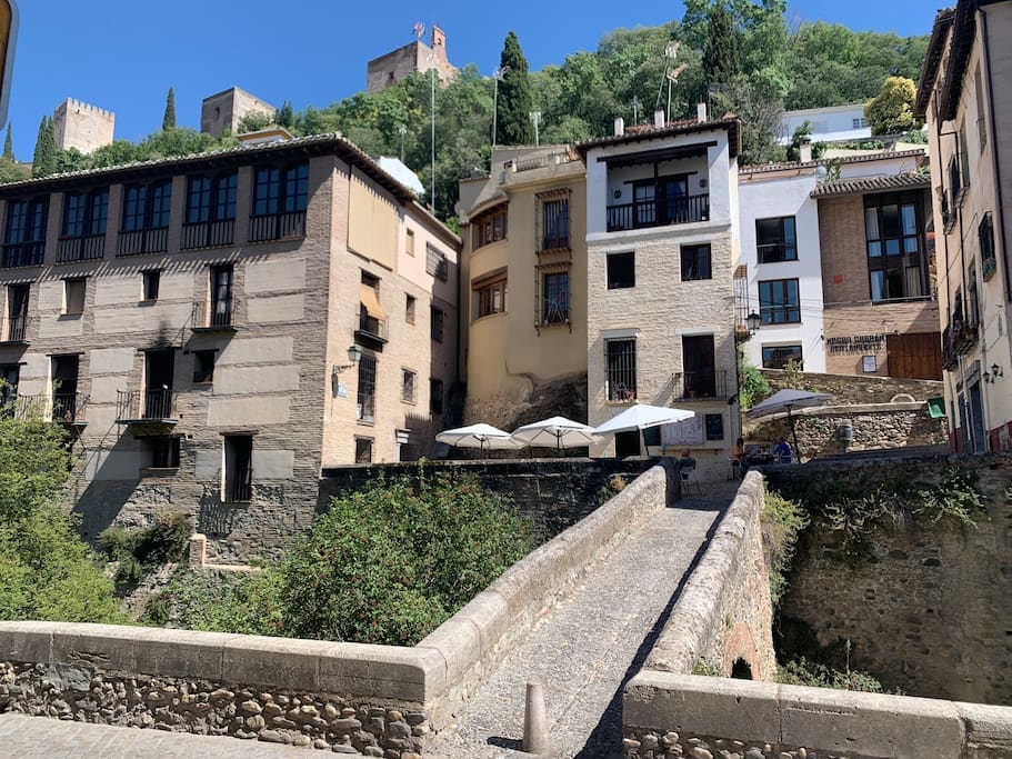 A series of charming buildings is set against a hillside, with trees and greenery surrounding the area. A narrow stone bridge leads to the structures, which feature a mix of architectural styles. Patios with umbrellas are visible, inviting outdoor relaxation.
