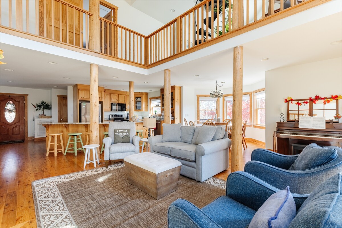 A spacious living area features warm wooden accents and tall ceilings, enhancing an open feel. Comfortable seating includes a gray sofa and two blue armchairs. A large area rug defines the space, with a coffee table centrally located. The kitchen and dining area are visible in the background.
