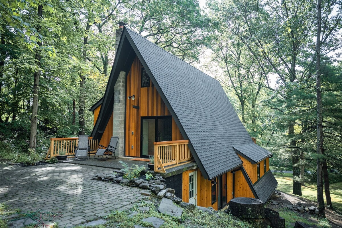 A striking A-frame structure is set among lush greenery, showcasing a blend of wood and stone exteriors. A spacious deck extends from the entrance, framed by a stone pathway, inviting a connection with the natural surroundings.