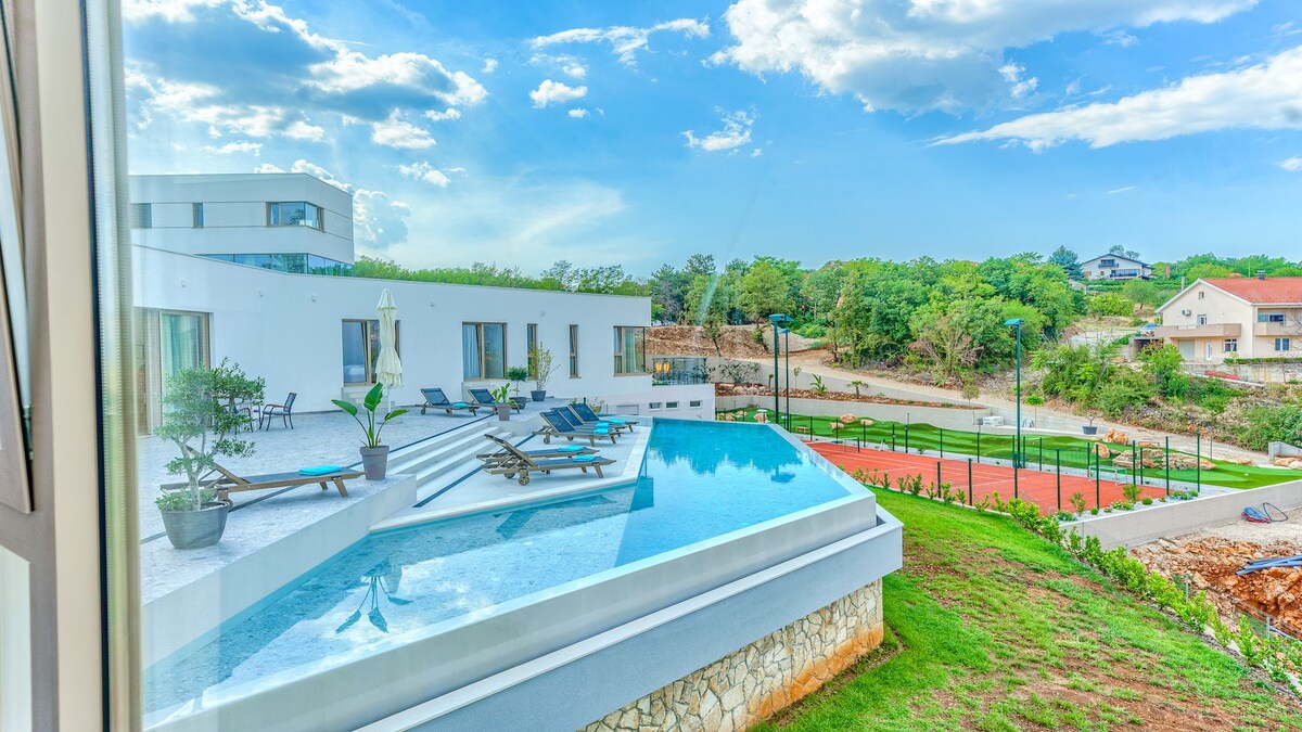 An infinity pool is visible, seamlessly extending from the villa's terrace, surrounded by sun loungers. Lush greenery provides a backdrop, complementing the pristine white architecture of the villa. A shared tennis court is situated in the distance, enhancing the outdoor recreational areas.