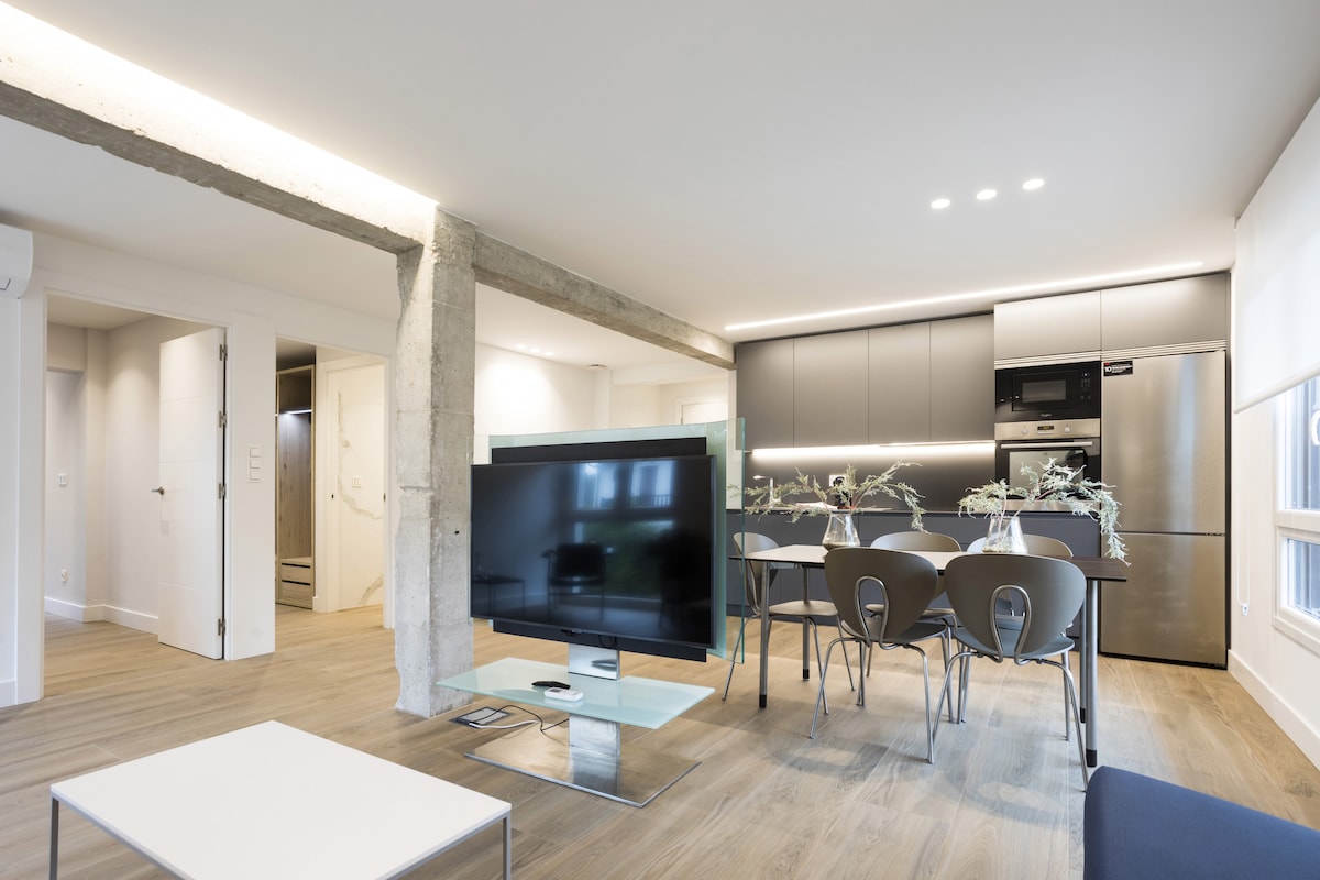 A modern living area features a sleek flat-screen television on a glass stand, facing a stylish dining table with four chairs. The kitchen area is visible in the background, showcasing contemporary appliances and cabinetry. Natural light is provided through large windows.