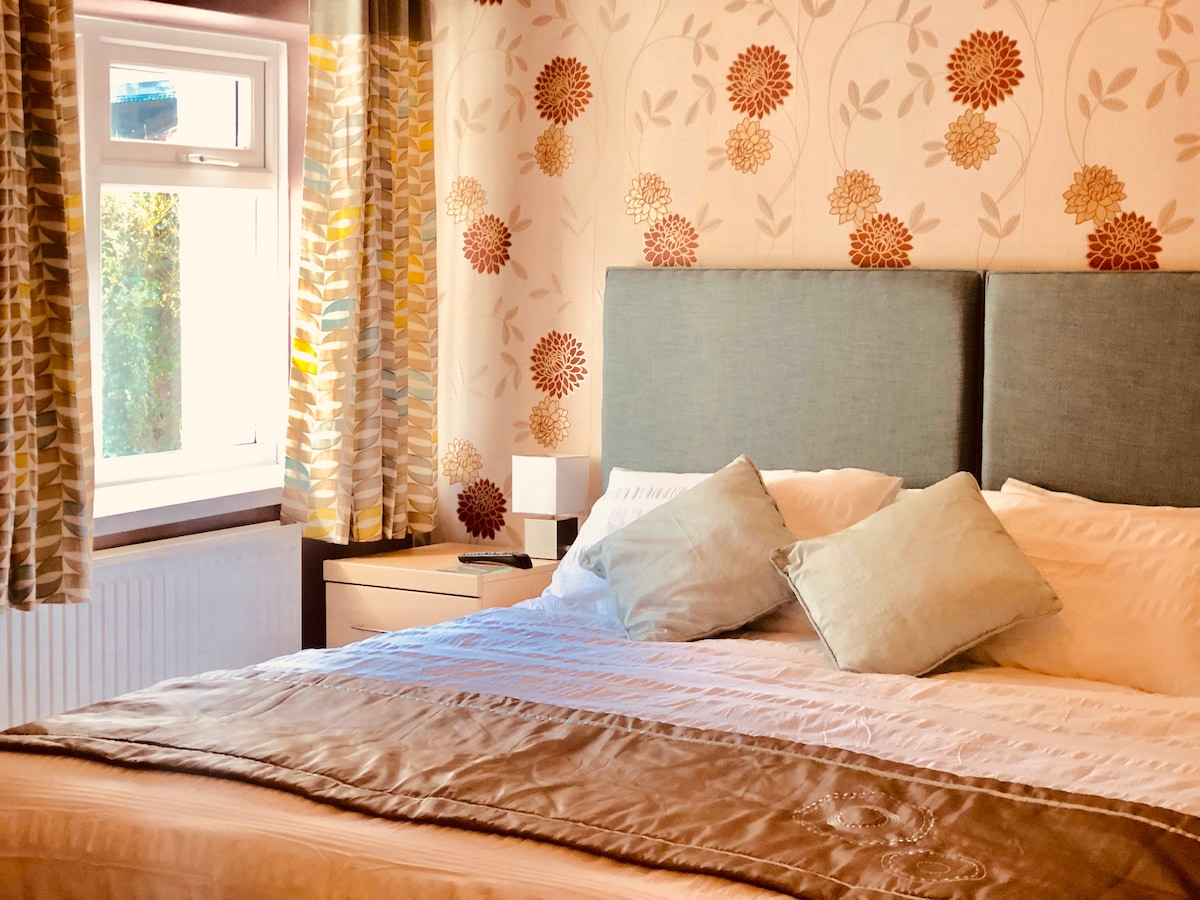 A well-appointed sleeping area features a spacious bed layered with soft linens and decorative pillows. Floral wallpaper adds a touch of warmth. Natural light enters through a window adorned with patterned curtains, enhancing the inviting ambiance of the room.