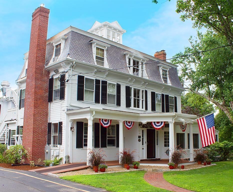 The historic Victorian Main house, built in the 1716 style, showcases classic architectural features, including dark shutters and a welcoming porch. American flags and decorative bunting enhance the exterior, while landscaped greenery and a brick pathway contribute to the property’s charming ambiance.
