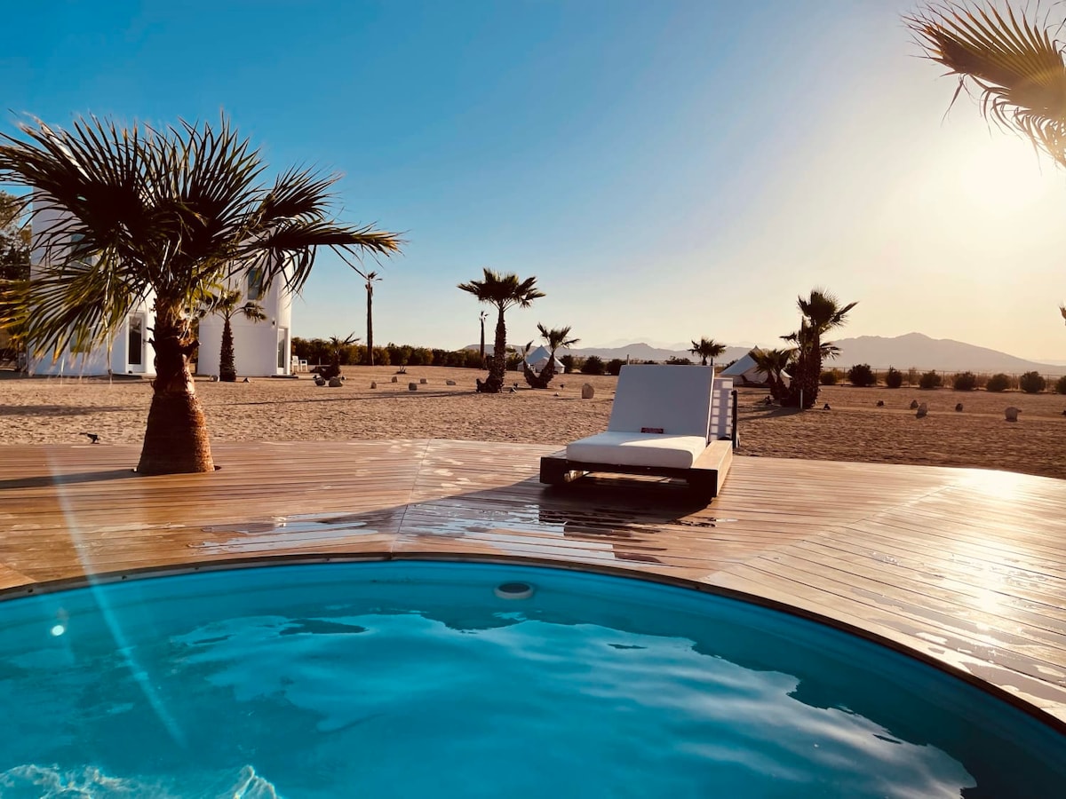 A circular cowboy pool is set on a wooden deck, reflecting the blue sky and sunlight. A single lounge chair is positioned beside the pool, with palm trees framing the scene. The expansive desert landscape and distant mountains provide a serene backdrop.