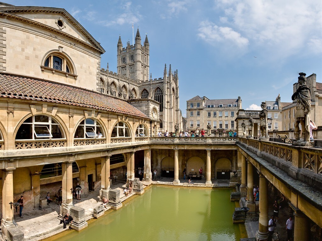 An expansive view of the historic Bath Roman Baths showcases arched columns surrounding the tranquil green pool. Stone sculptures and visitors can be seen along the walkways, with the impressive Abbey in the background under a clear blue sky.